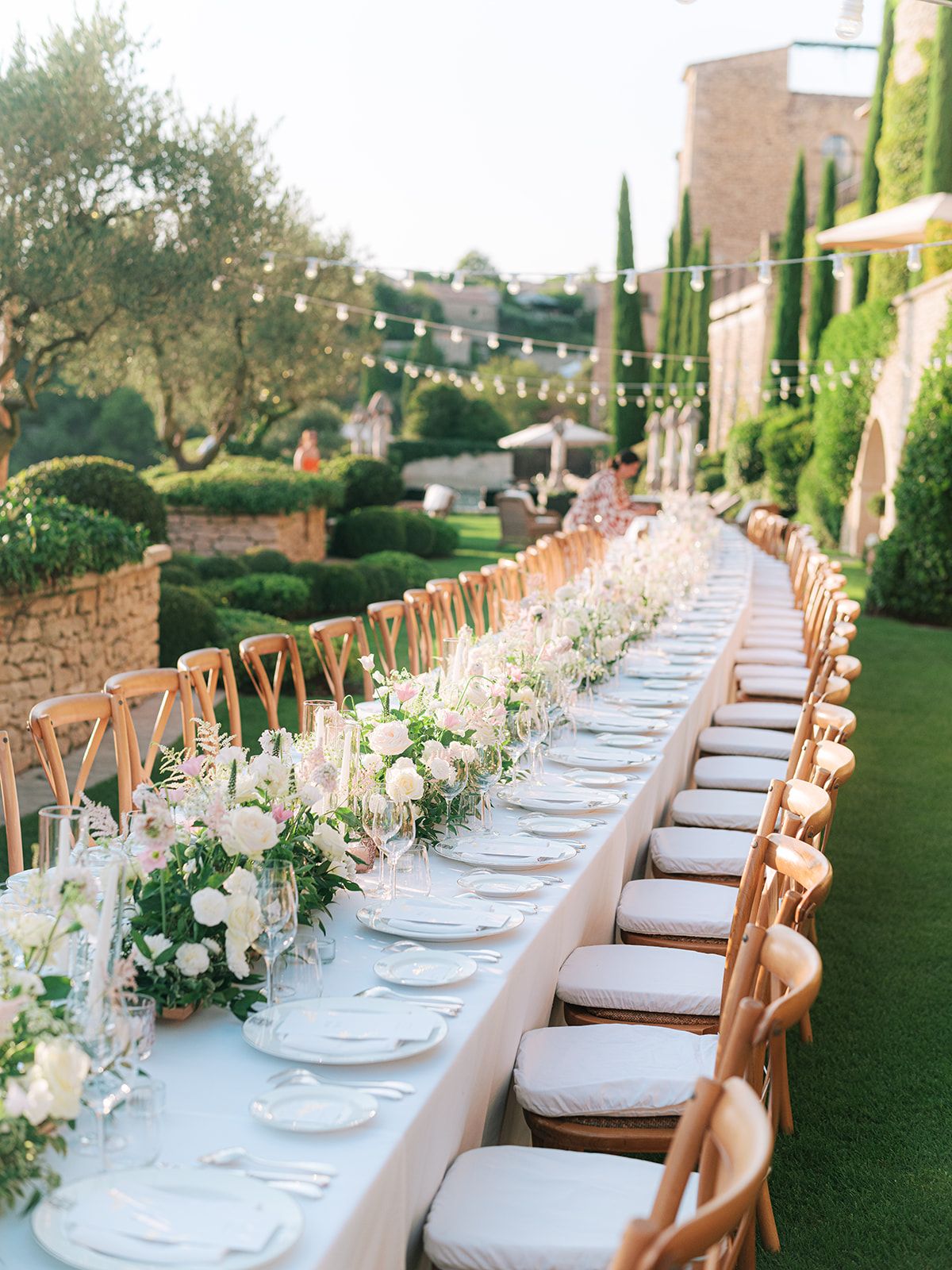 Long banquet table with white and blush floral runner, cross-back chairs, and bistro lights in chateau garden