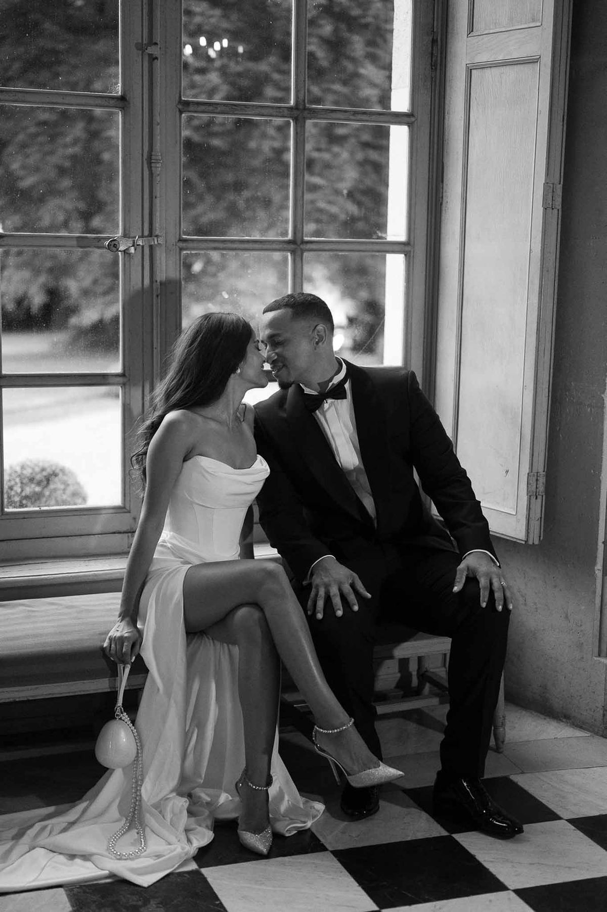 Black-and-white portrait of bride and groom seated by French windows on checkerboard tile floor in chateau interior