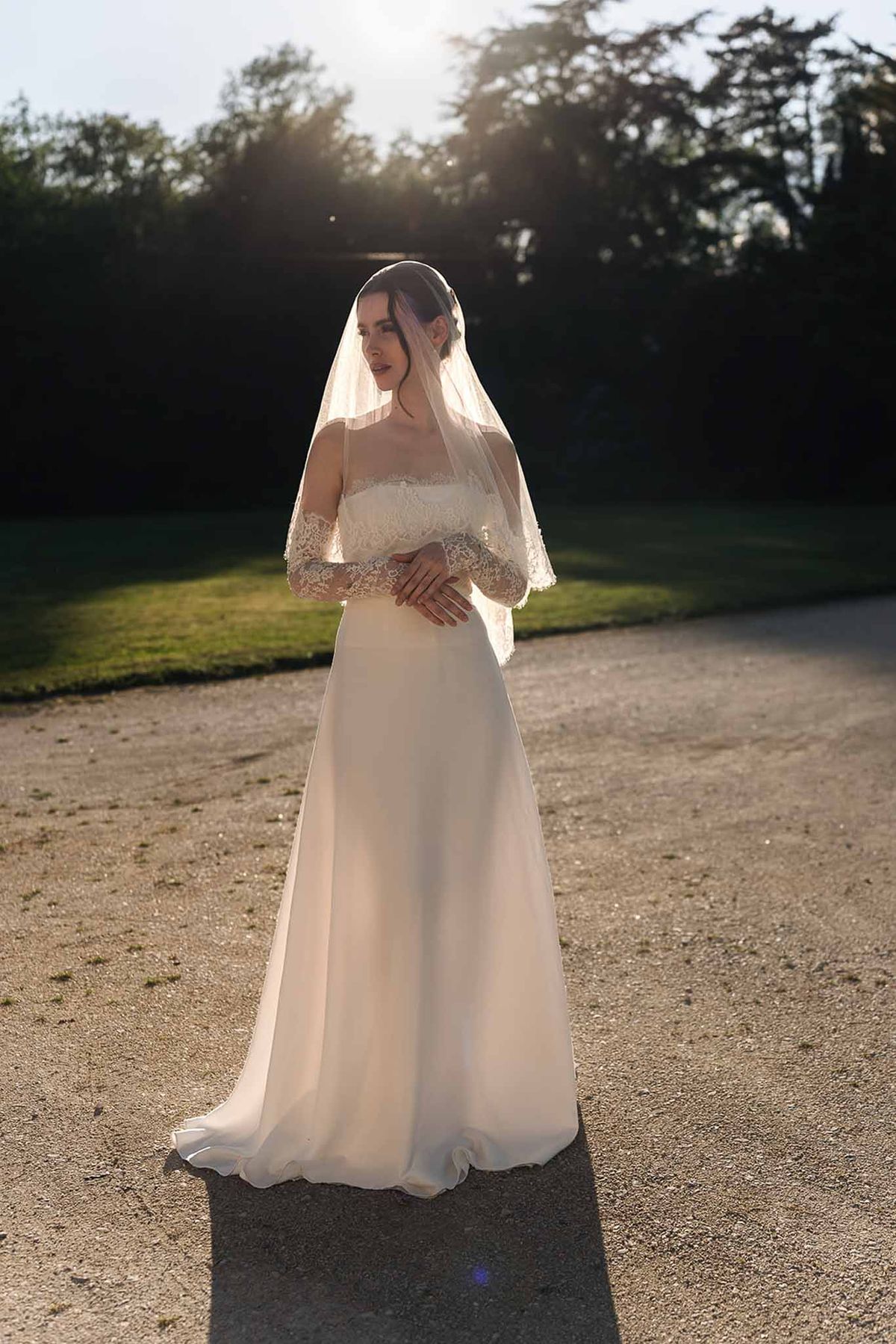 Bride in ivory lace off-the-shoulder gown and lace-trimmed veil backlit by golden sunset on a gravel path