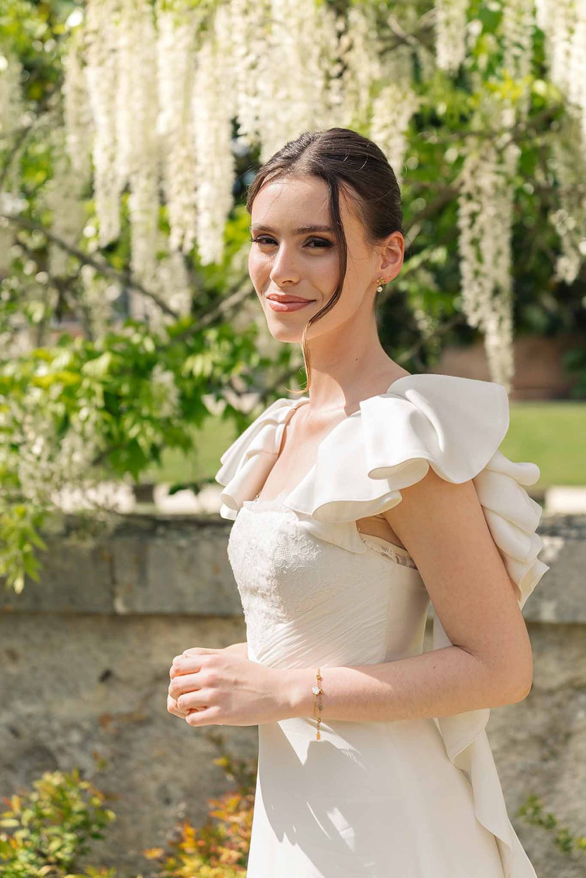 Bride in ivory gown with structured ruffle sleeves and lace bodice, hair in sleek updo, white wisteria in background