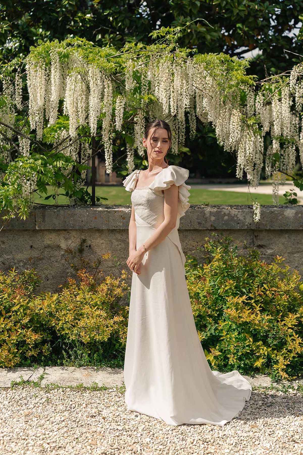 Bride in ivory A-line gown with ruffled cap sleeve standing before wisteria-draped stone balustrade in chateau garden