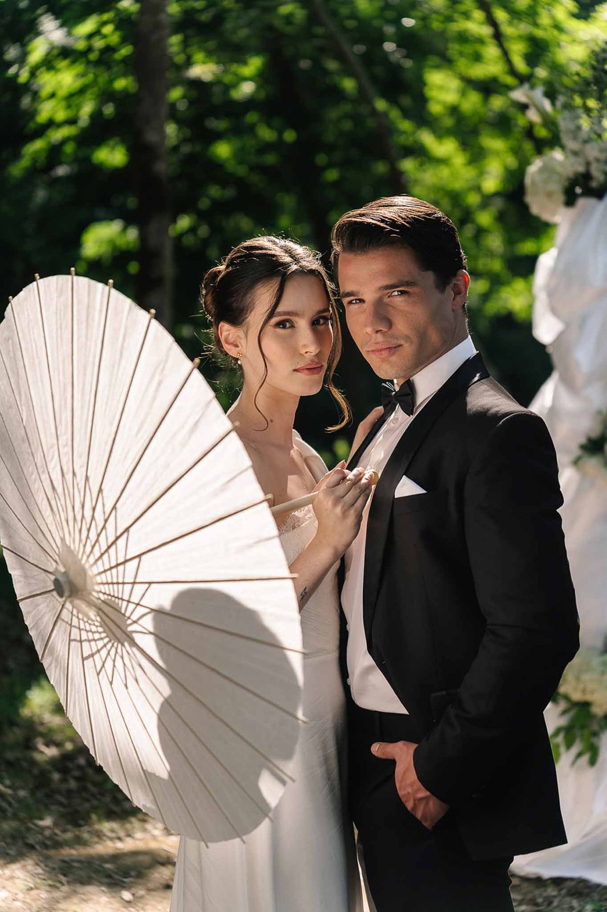 Couple in wooded garden bride in white slip gown with paper parasol groom in black tuxedo editorial pose