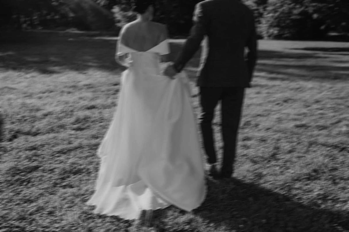Motion-blurred couple walks arm-in-arm across lawn in off-shoulder gown and dark suit in B&W