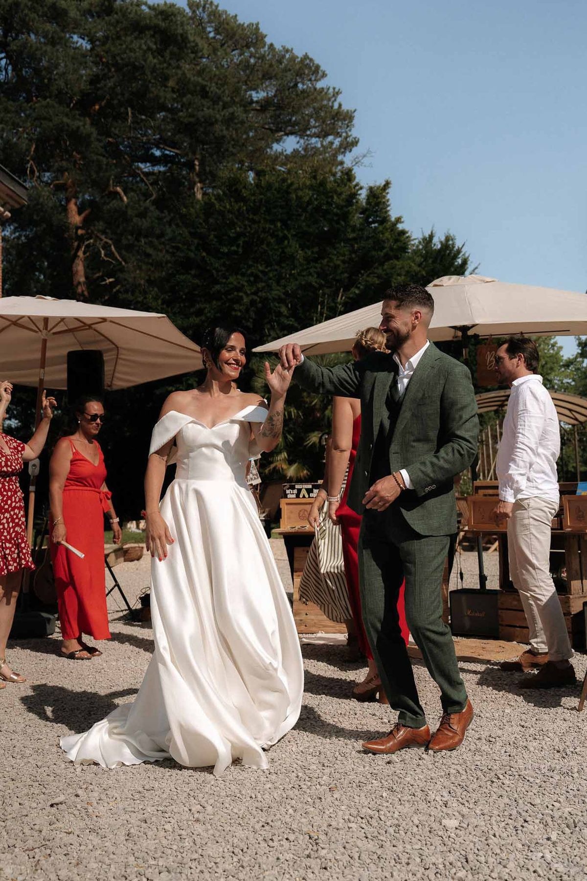 Bride in white off-the-shoulder ballgown spinning with groom in green tweed suit on gravel courtyard