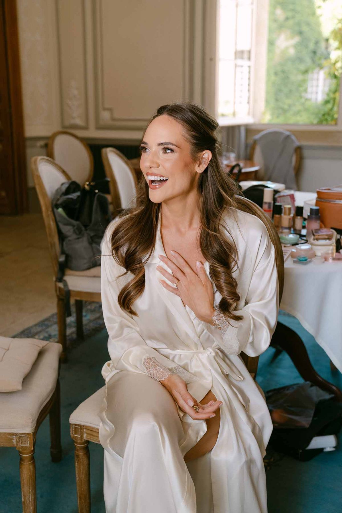 Bride in cream satin robe with voluminous waves laughing on French chair in panelled chateau room