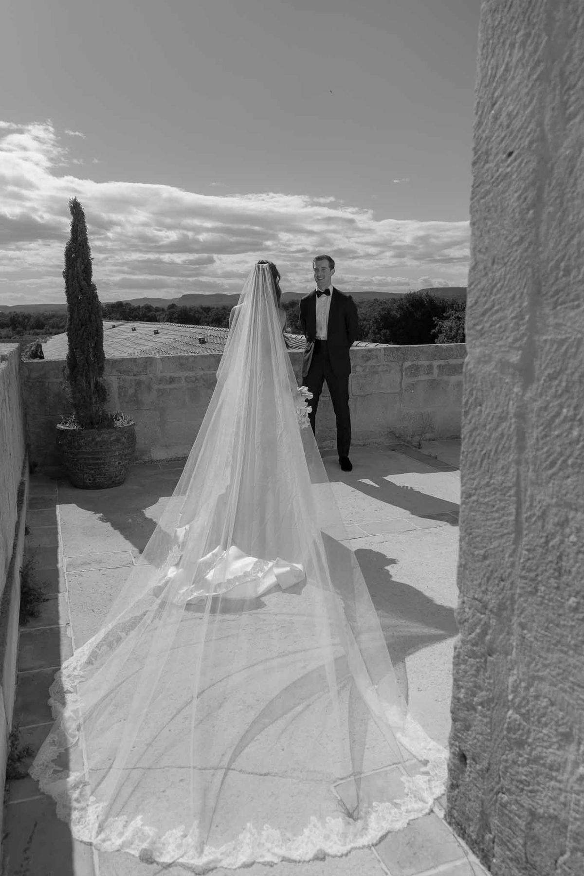Black and white groom smiling as bride faces away with lace-trimmed veil on chateau terrace with hills