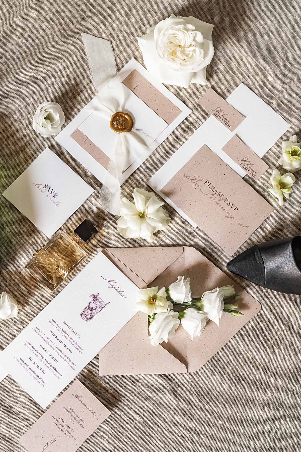 Stationery suite with gold wax seal, ivory ribbon, burgundy script, ivory roses, and perfume bottle on linen