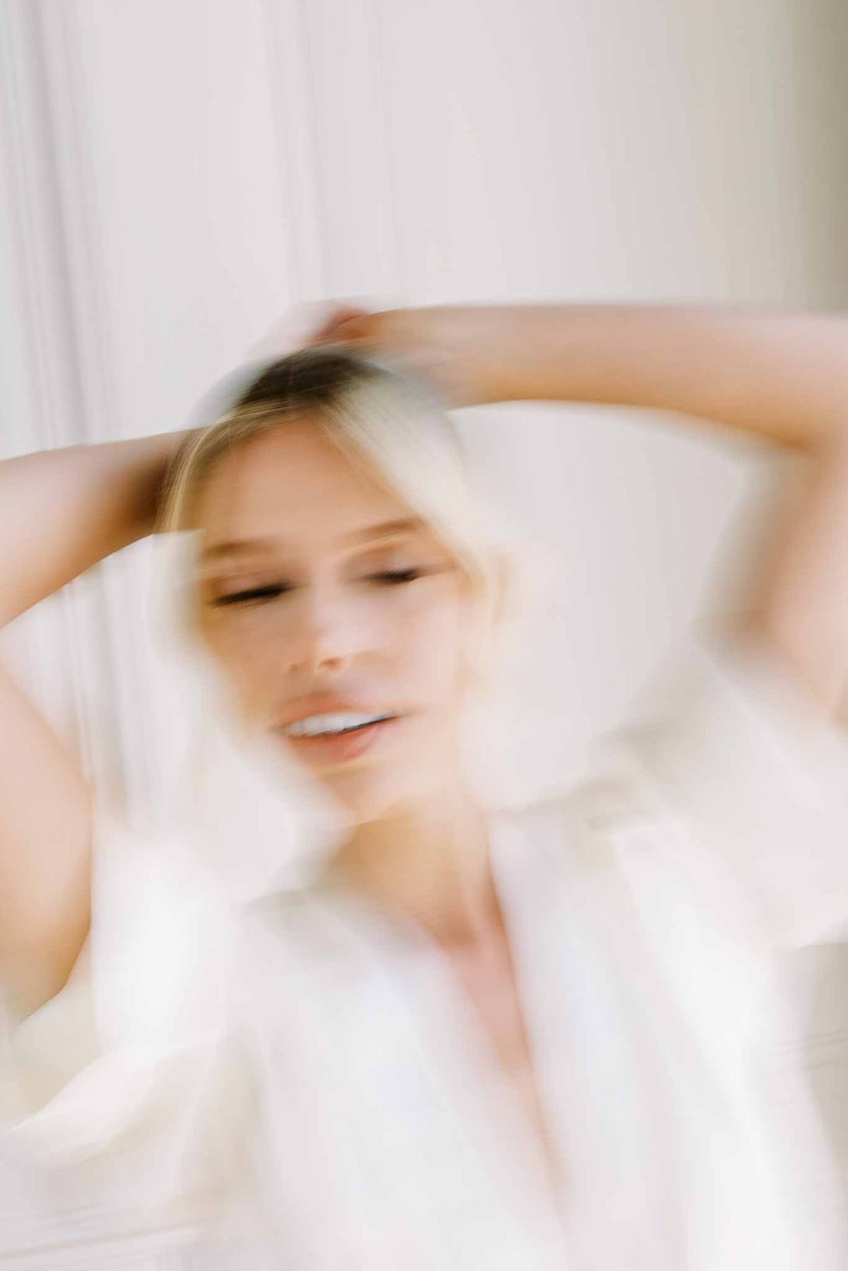Motion-blurred artistic portrait of bride adjusting hair in ivory satin robe during getting ready