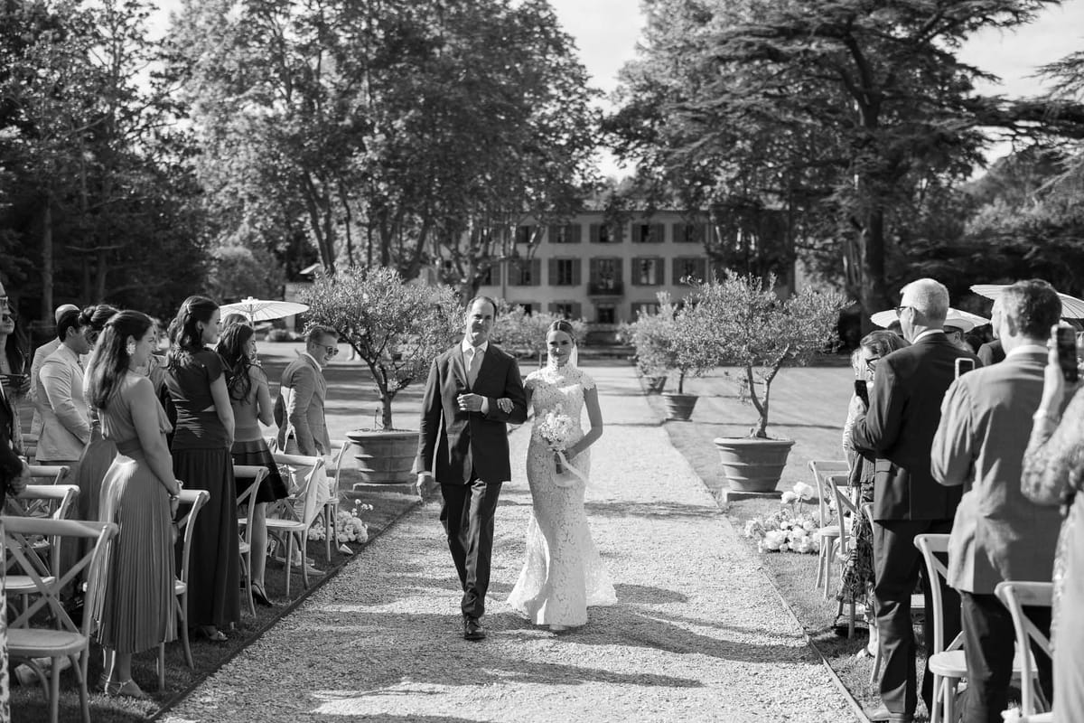 Black and white bride in lace gown escorted down aisle lined with potted olive trees before classical manor house
