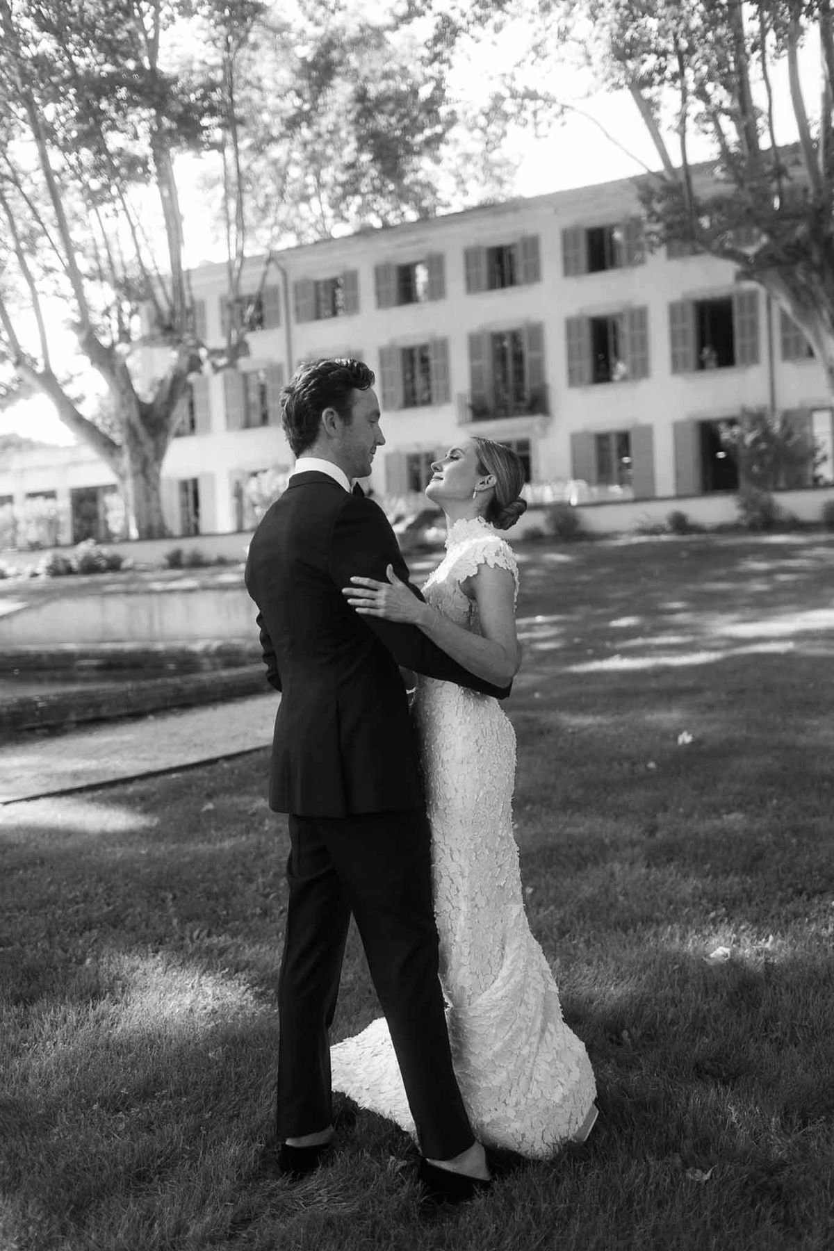 Black and white full-length portrait of bride in fitted lace gown embracing groom on chateau lawn
