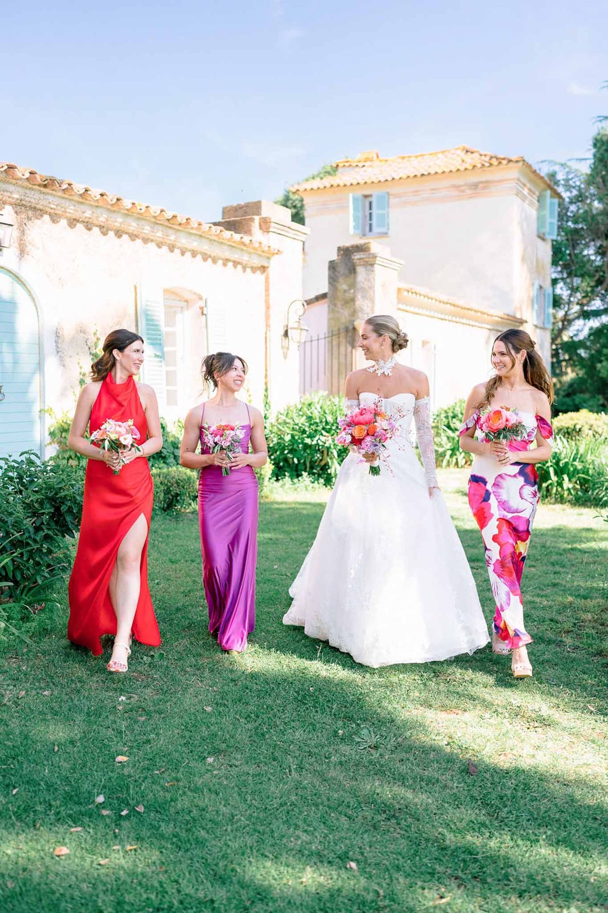 Bride in white ball gown walking with three bridesmaids in red, purple, and floral outfits on lawn