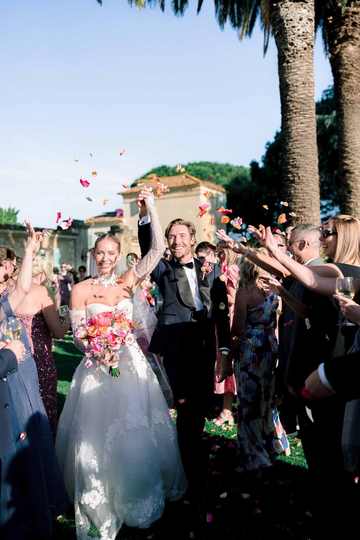 Couple raises joined hands in hot pink and coral petal toss with bride's vibrant peony bouquet at estate