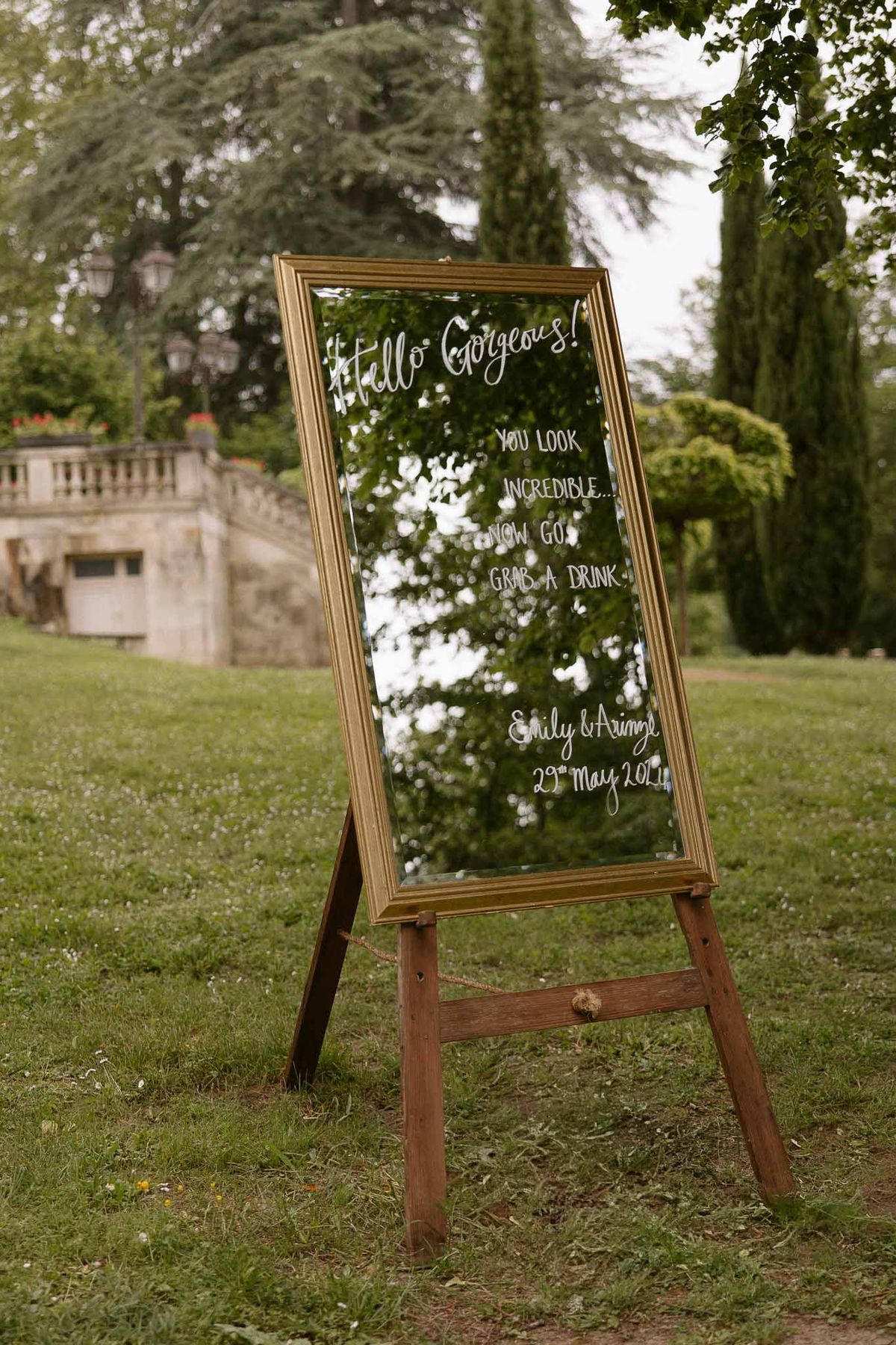 Gold-framed welcome mirror sign with white calligraphy on lawn easel with stone balustrade behind