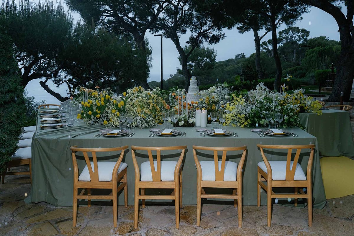Dusk reception table with sage green linen, yellow tulips, white orchids, and tiered cake flanked by pillar candles on ter...
