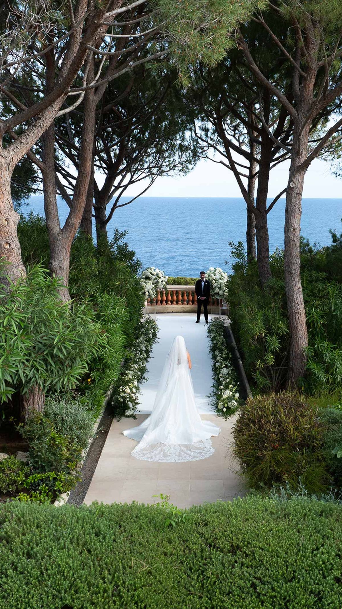 Elevated view of bride walking white aisle toward groom on coastal terrace with hydrangea arrangements