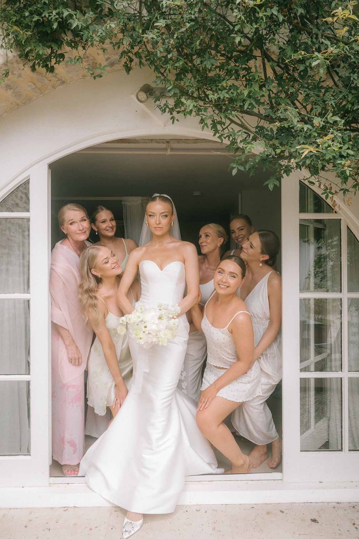 Bride in strapless satin gown with six bridesmaids in pink robes posed in chateau arched doorway