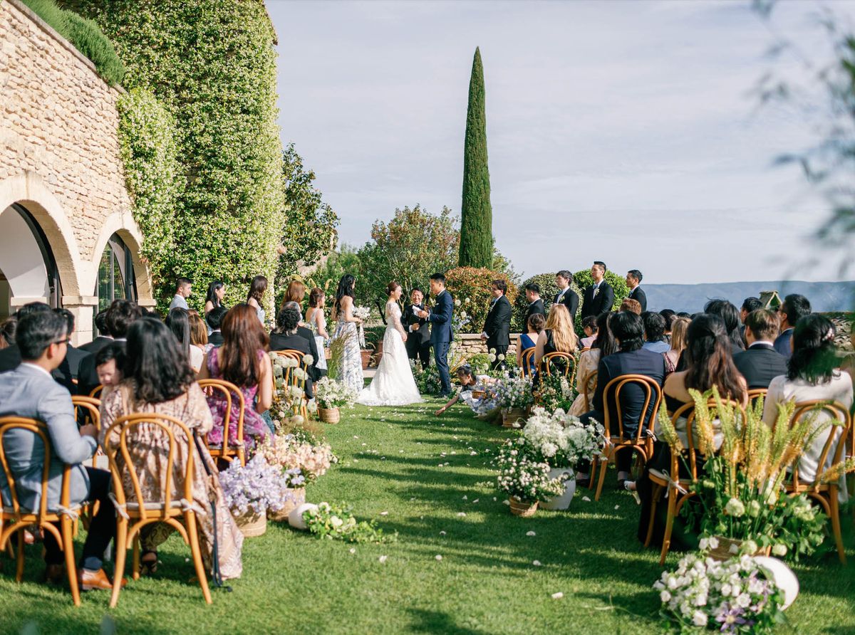 Ceremony beside ivy-covered building with wicker basket florals of allium and lavender on grass aisle