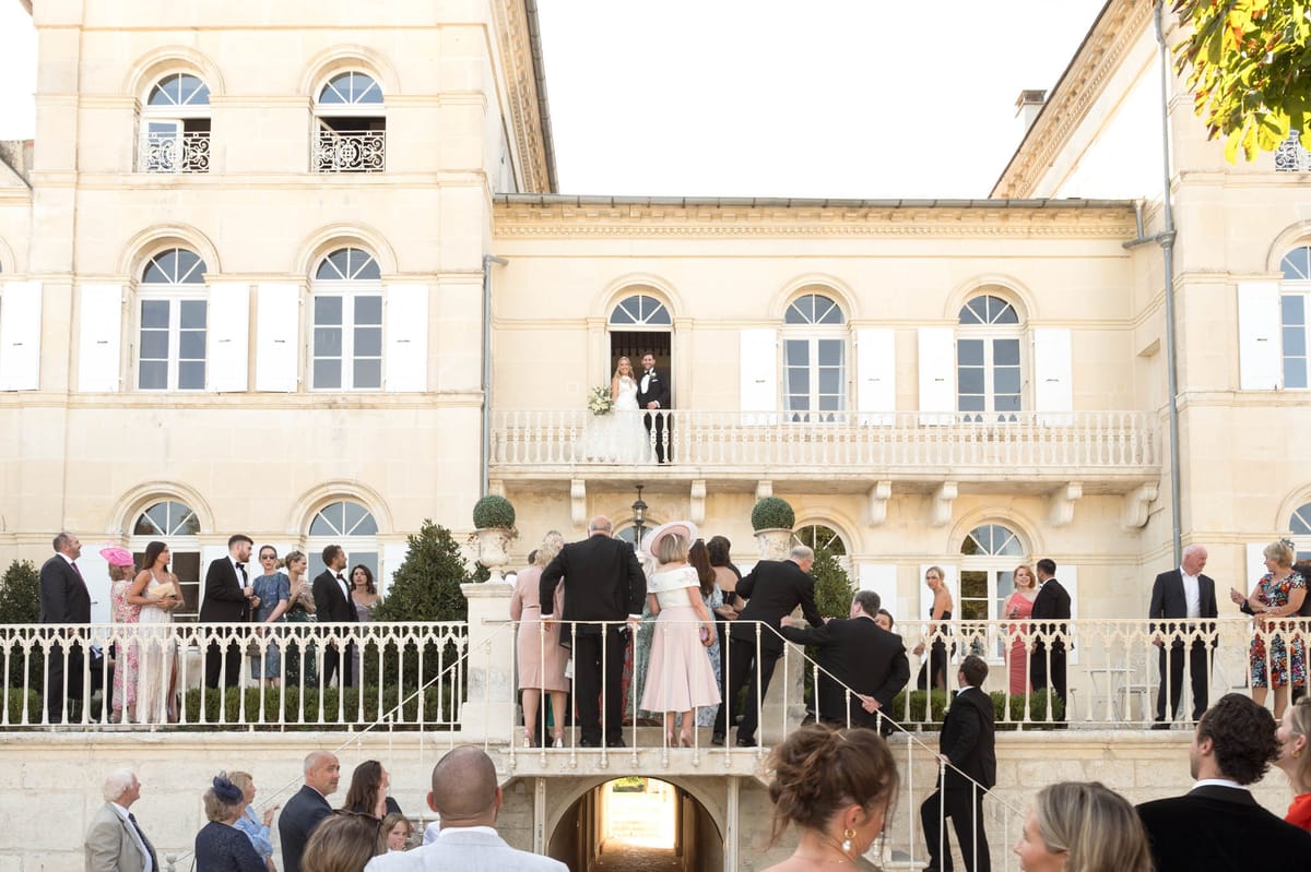 Couple on chateau balcony overlooking 30 guests gathered on terrace during cocktail hour in late afternoon light