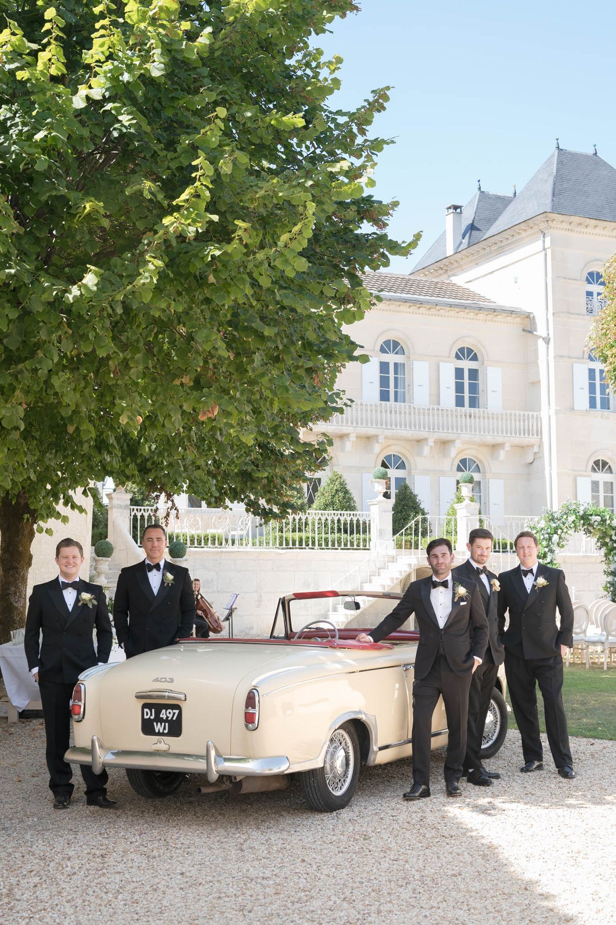 Five men in black tuxedos posing beside vintage cream Peugeot convertible at chateau grounds
