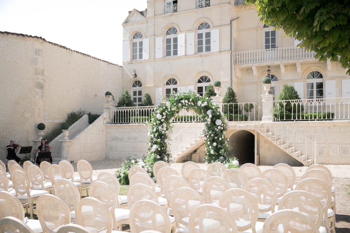 Outdoor ceremony setup with white rose arch, Louis XVI chairs, and string duo in a French chateau courtyard