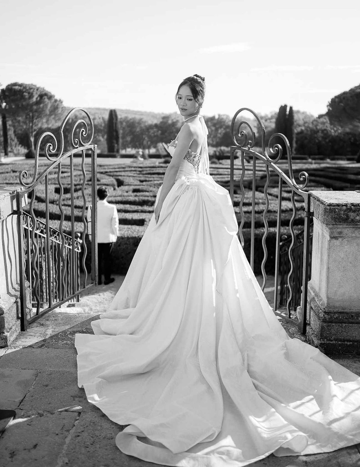 Bride in halter-neck ball gown at wrought-iron gate overlooking parterre gardens at Château de la Gaude