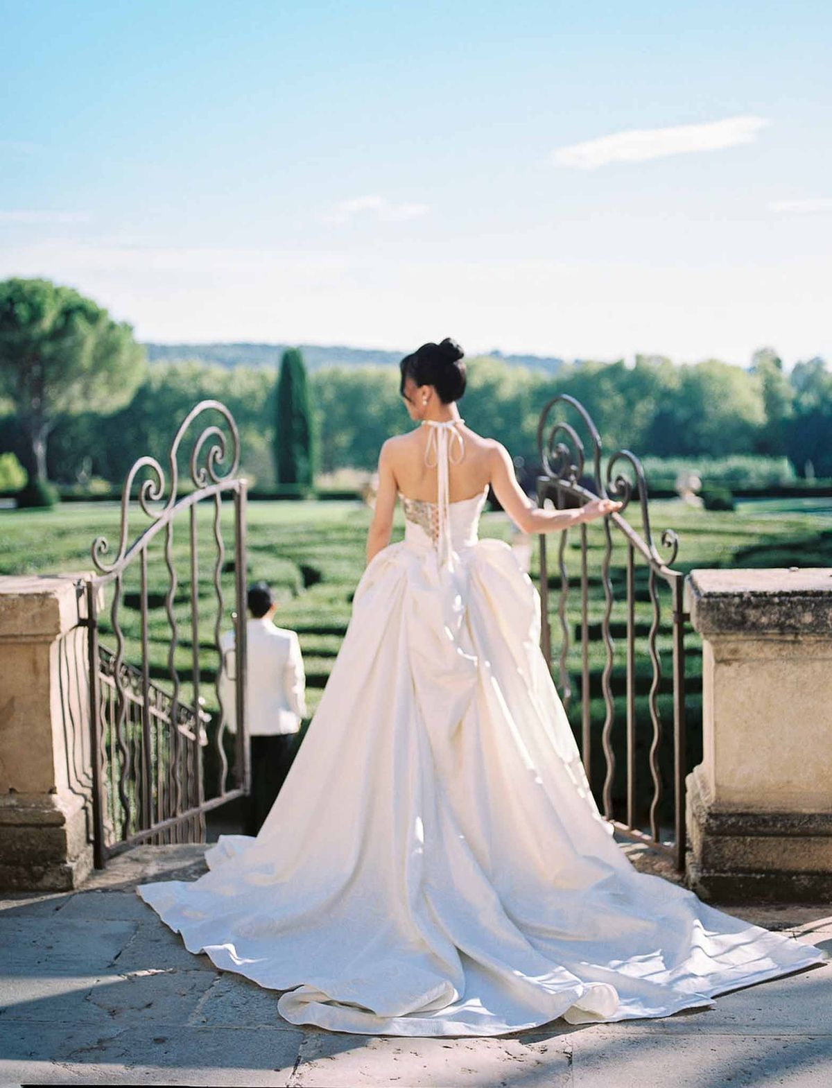 Back view of bride with long train at iron gate overlooking formal gardens at Château de la Gaude