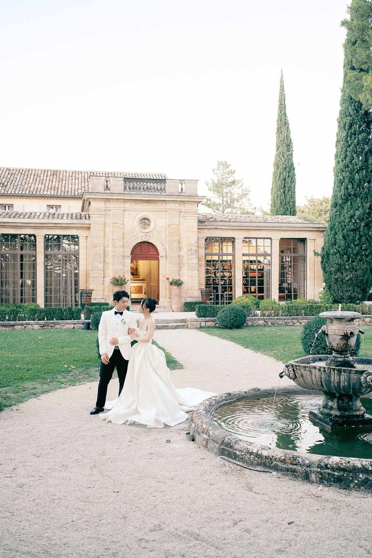 Bride and groom walking by stone fountain with orangery at Château de la Gaude