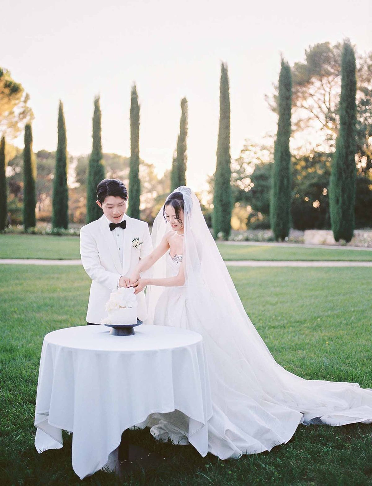 Bride and groom cutting wedding cake outdoors with cypress trees at sunset at Château de la Gaude