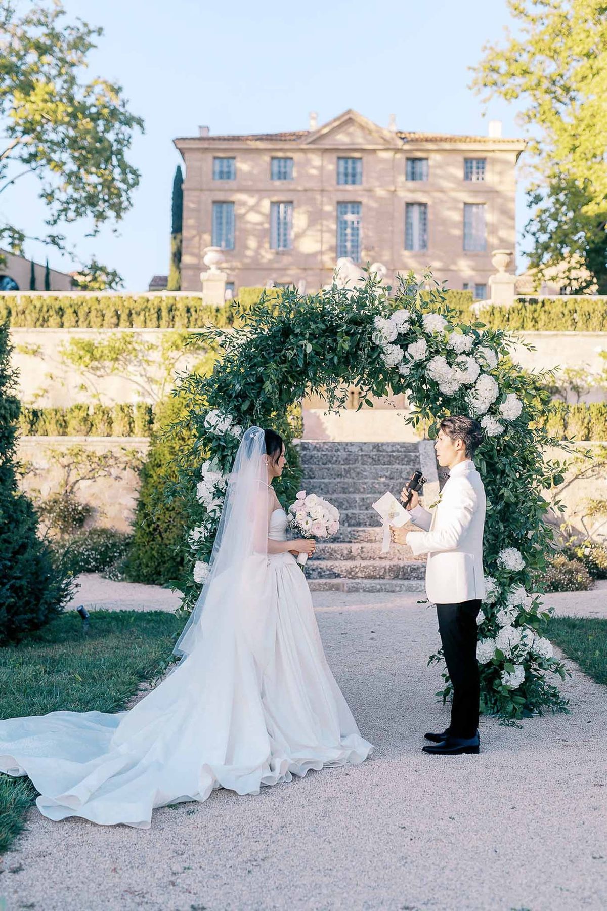 Couple exchanging vows under greenery arch with château façade behind at Château de la Gaude