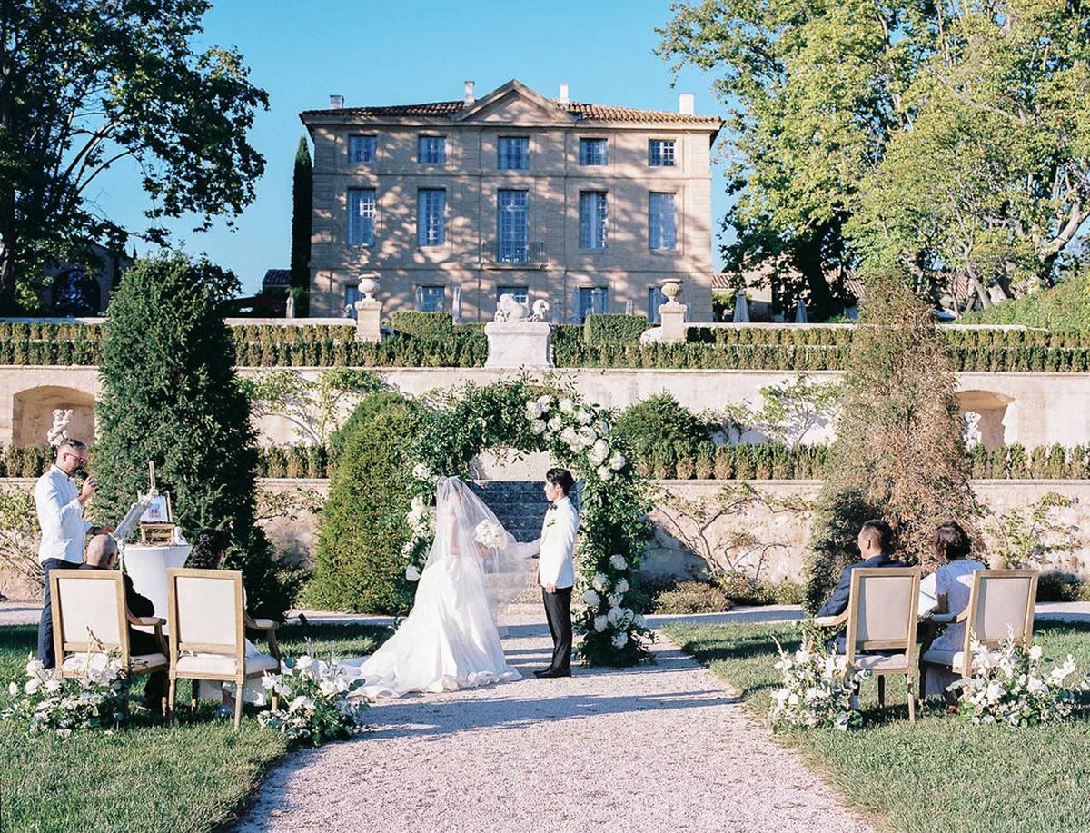 Wide view of outdoor ceremony with guests, greenery arch and château at Château de la Gaude