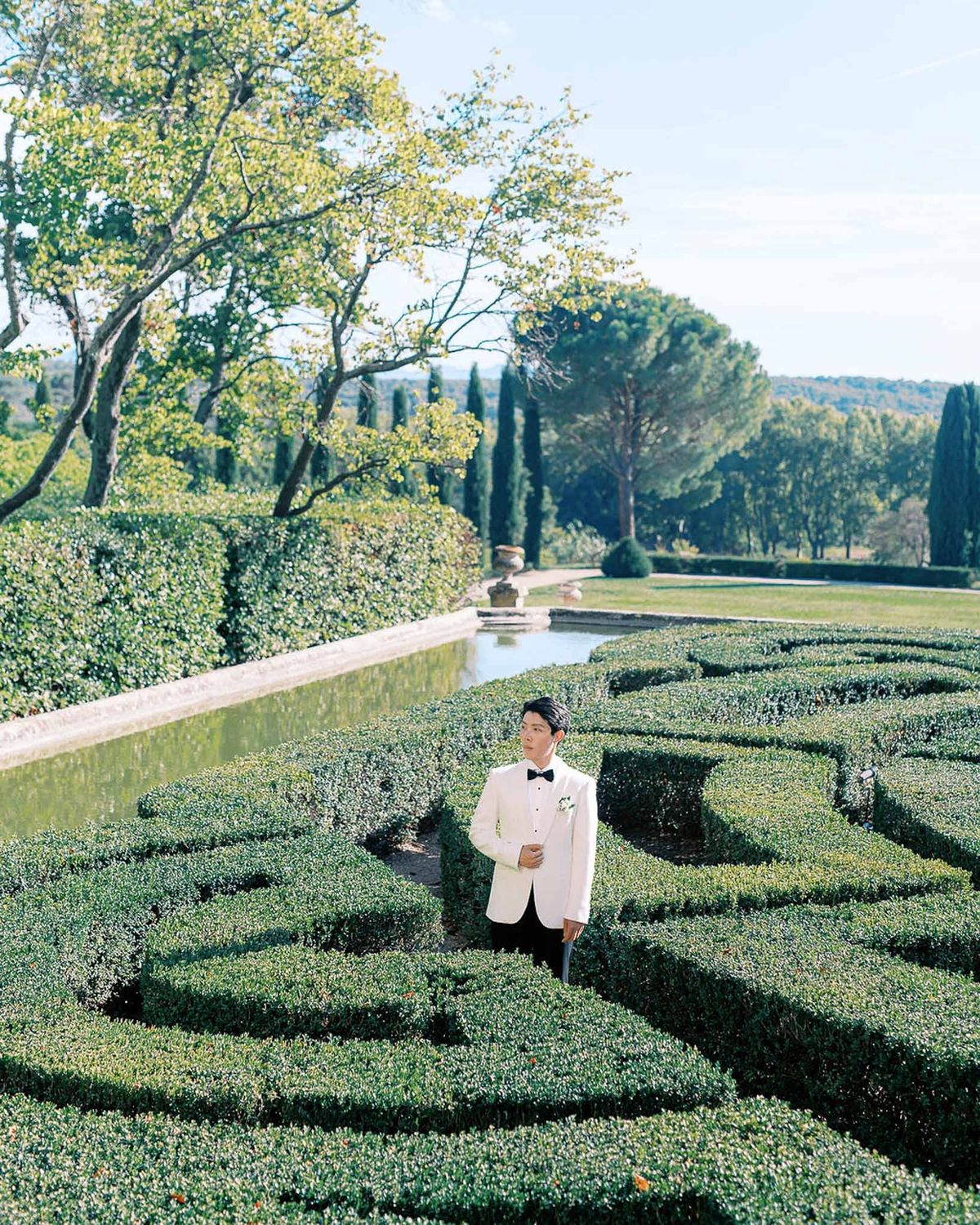 Groom walking through parterre maze garden in white dinner jacket at Château de la Gaude