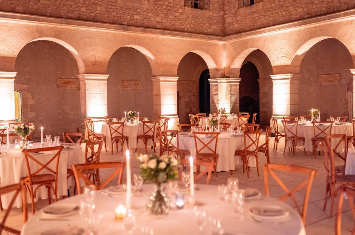 Round reception tables with white linens and cross-back chairs set inside a stone abbey cloister with amber uplighting