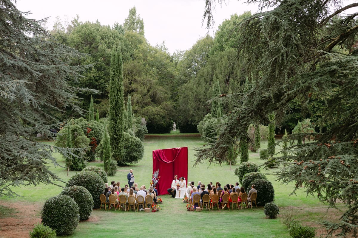 Rooa and Mark's outdoor garden wedding ceremony at Domaine de Perrotin, set before a raspberry-red draped backdrop with asymmetric colourful florals along a cypress-lined lawn