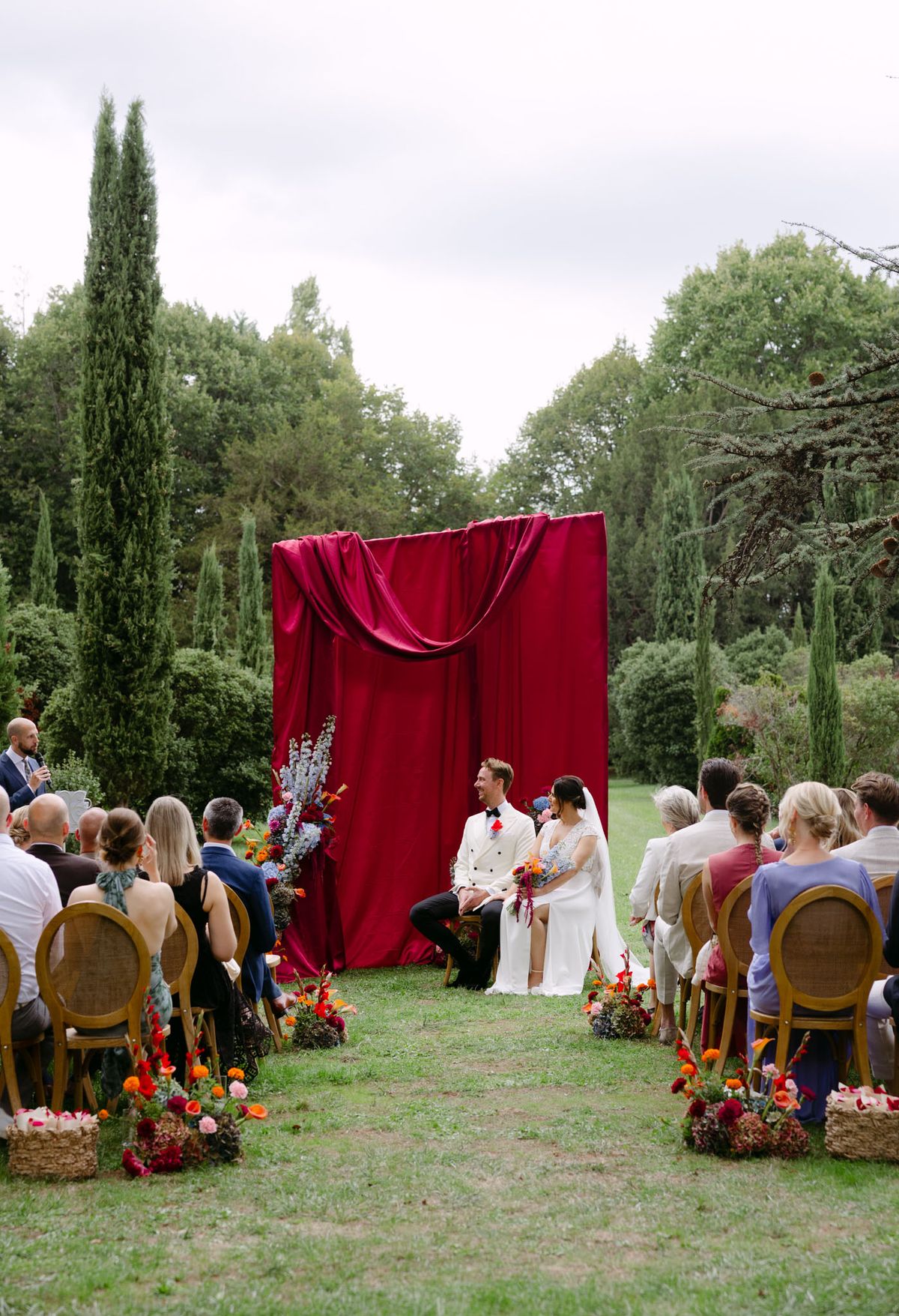 Rooa and Mark's outdoor garden wedding ceremony at Domaine de Perrotin, set before a raspberry-red draped backdrop with asymmetric colourful florals along a cypress-lined lawn