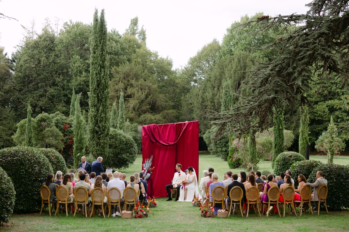 Rooa and Mark's outdoor garden wedding ceremony at Domaine de Perrotin, set before a raspberry-red draped backdrop with asymmetric colourful florals along a cypress-lined lawn