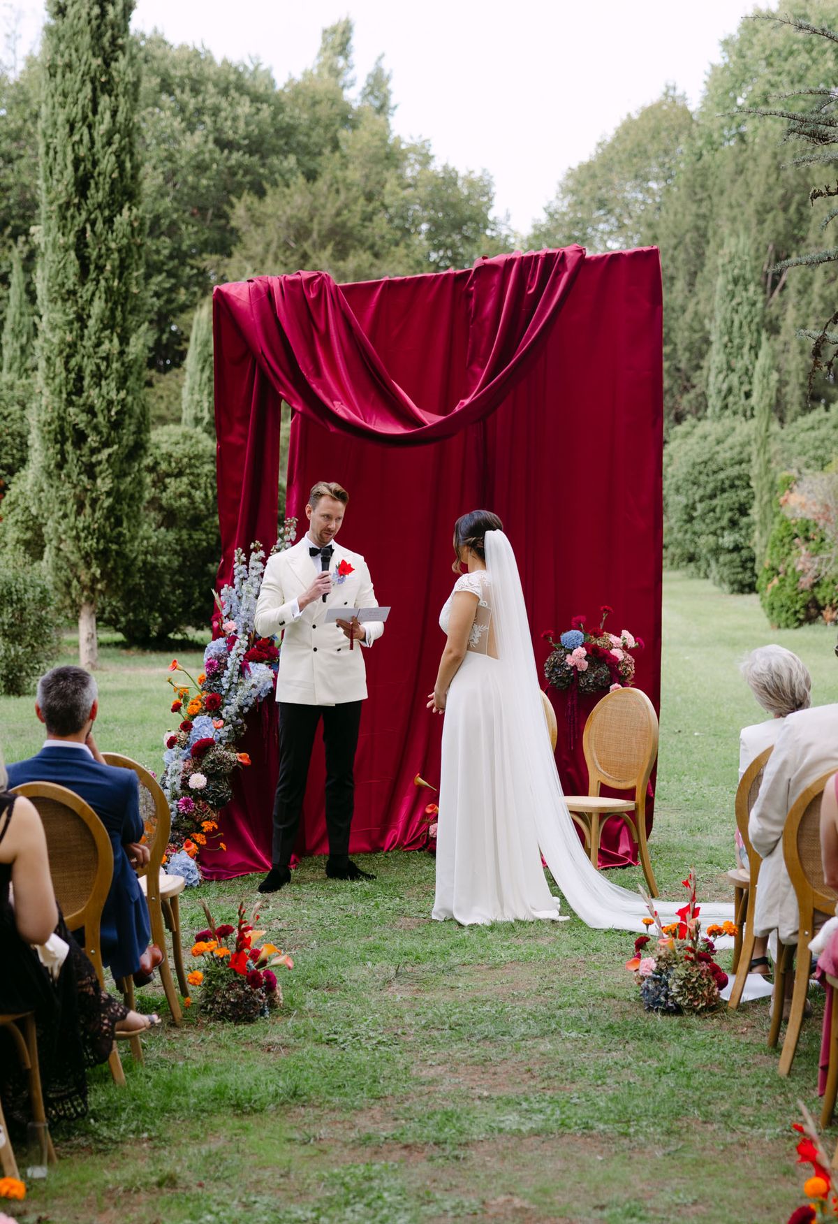 Rooa and Mark's outdoor garden wedding ceremony at Domaine de Perrotin, set before a raspberry-red draped backdrop with asymmetric colourful florals along a cypress-lined lawn