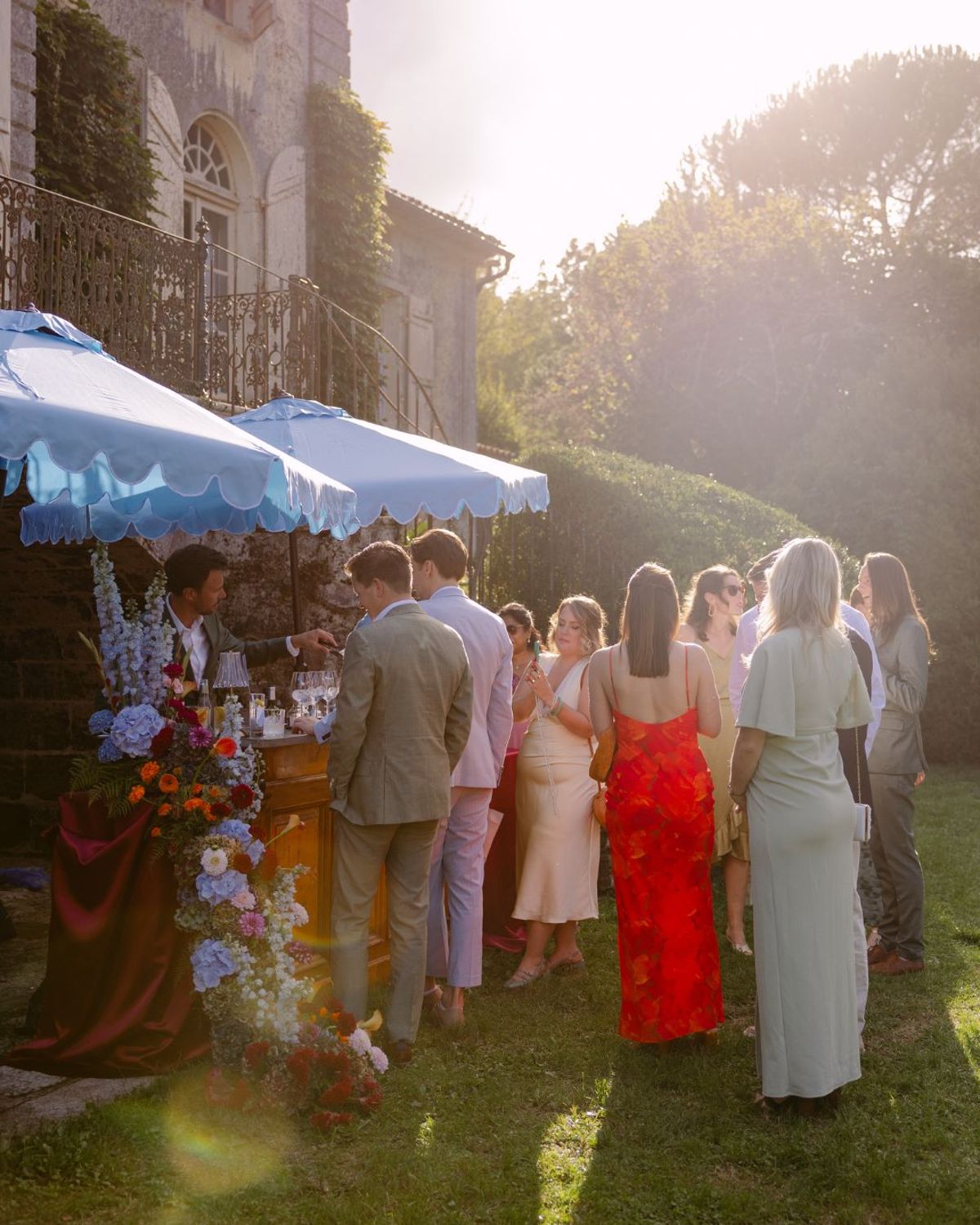 Guests gather for golden-hour welcome drinks at the flower-dressed bar under sky-blue parasols in the gardens of Domaine de Perrotin, for Rooa and Mark's wedding