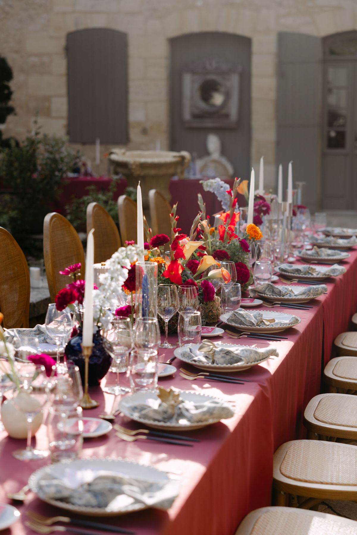 Outdoor reception in the stone patio courtyard at Domaine de Perrotin, with long tables dressed in raspberry-red linen, gold cane chairs and colourful florals for Rooa and Mark's wedding