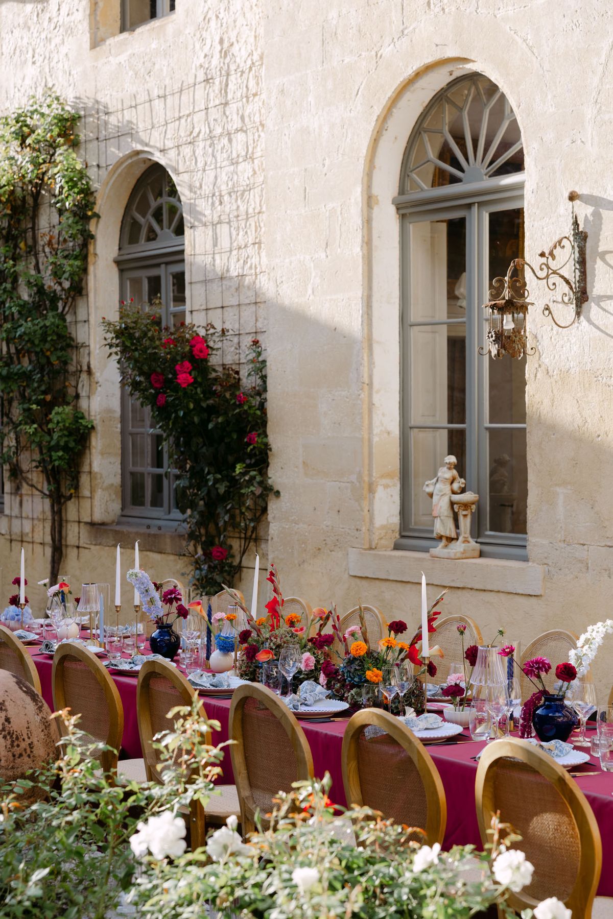Outdoor reception in the stone patio courtyard at Domaine de Perrotin, with long tables dressed in raspberry-red linen, gold cane chairs and colourful florals for Rooa and Mark's wedding