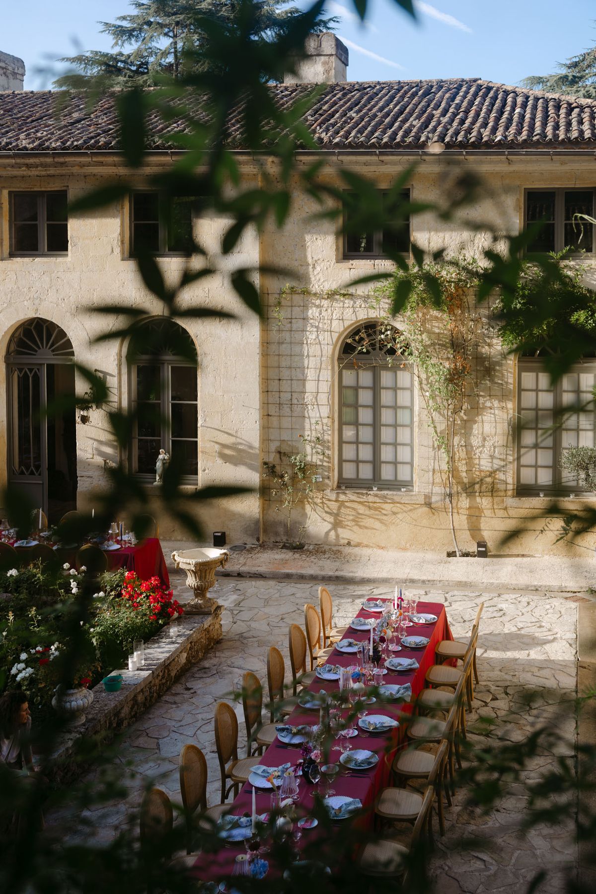 Outdoor reception in the stone patio courtyard at Domaine de Perrotin, with long tables dressed in raspberry-red linen, gold cane chairs and colourful florals for Rooa and Mark's wedding