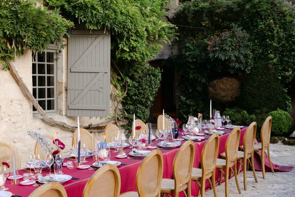 Outdoor reception in the stone patio courtyard at Domaine de Perrotin, with long tables dressed in raspberry-red linen, gold cane chairs and colourful florals for Rooa and Mark's wedding