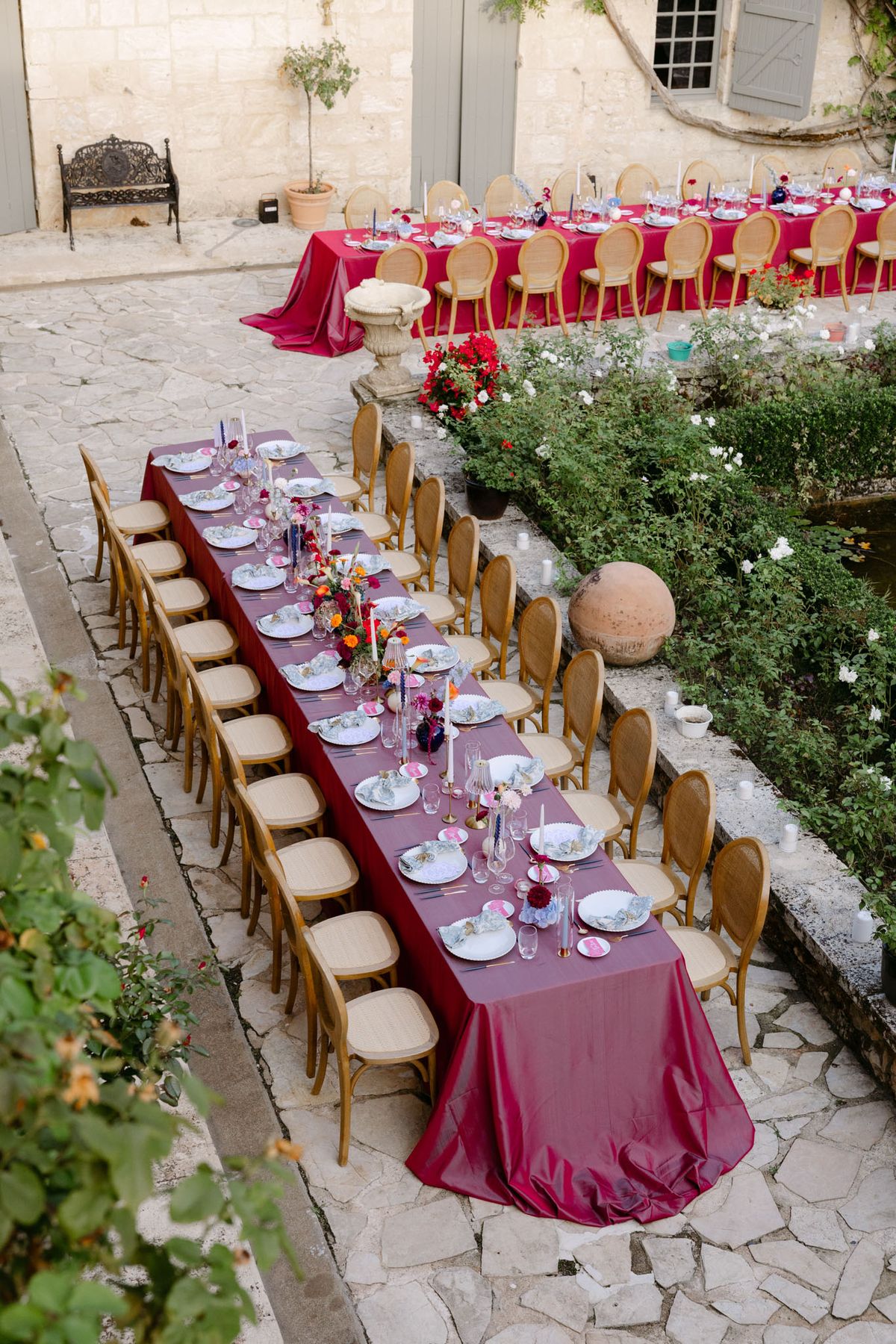 Outdoor reception in the stone patio courtyard at Domaine de Perrotin, with long tables dressed in raspberry-red linen, gold cane chairs and colourful florals for Rooa and Mark's wedding