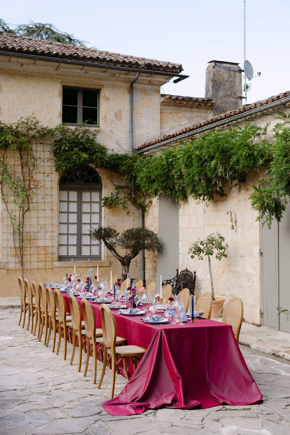 Outdoor reception in the stone patio courtyard at Domaine de Perrotin, with long tables dressed in raspberry-red linen, gold cane chairs and colourful florals for Rooa and Mark's wedding