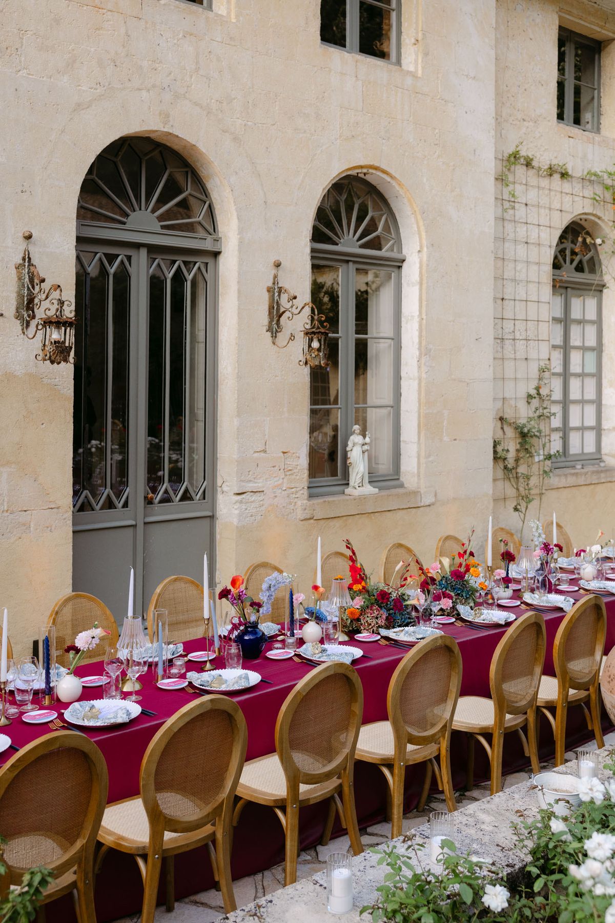 Outdoor reception in the stone patio courtyard at Domaine de Perrotin, with long tables dressed in raspberry-red linen, gold cane chairs and colourful florals for Rooa and Mark's wedding
