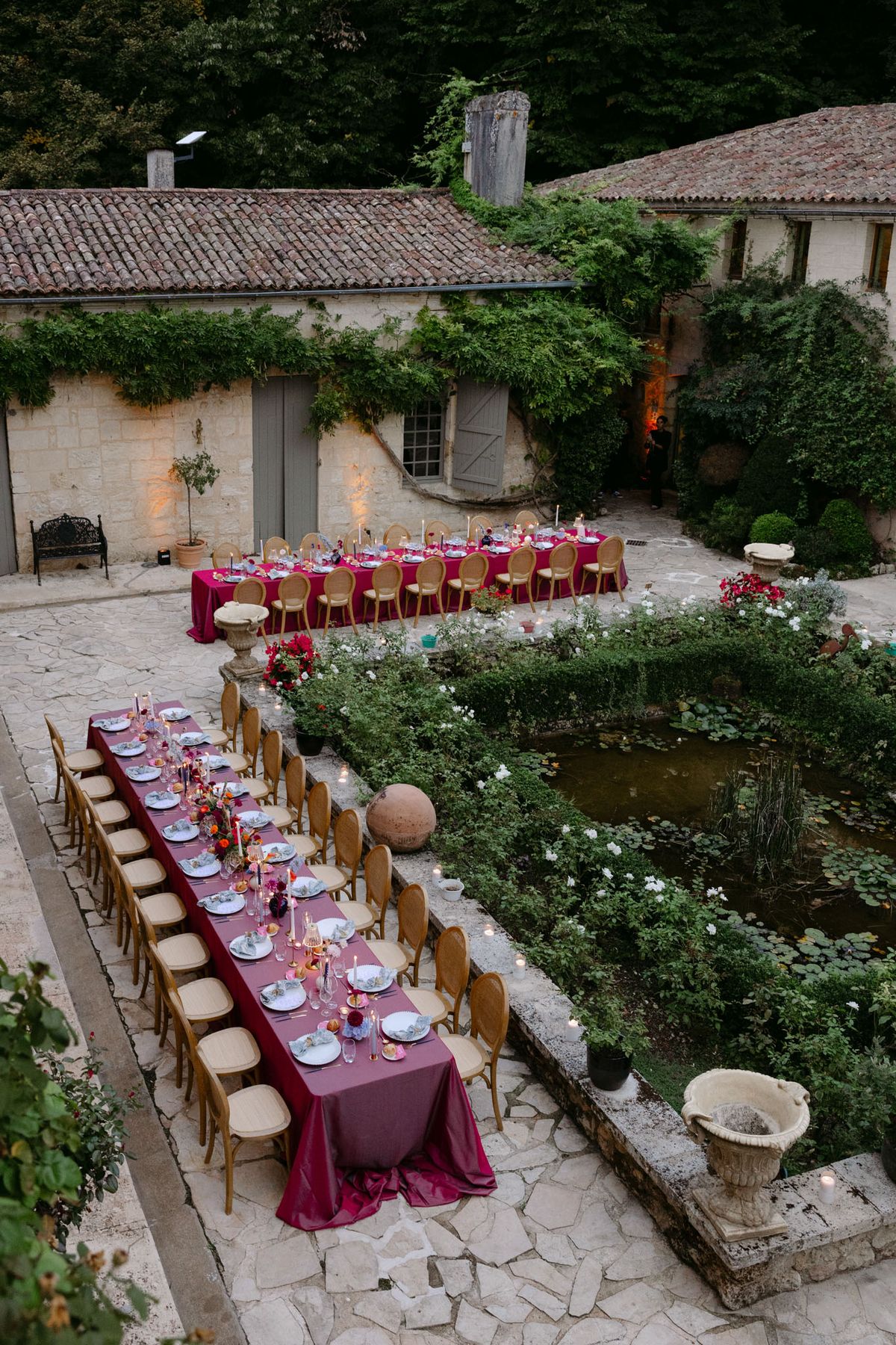 Outdoor reception in the stone patio courtyard at Domaine de Perrotin, with long tables dressed in raspberry-red linen, gold cane chairs and colourful florals for Rooa and Mark's wedding