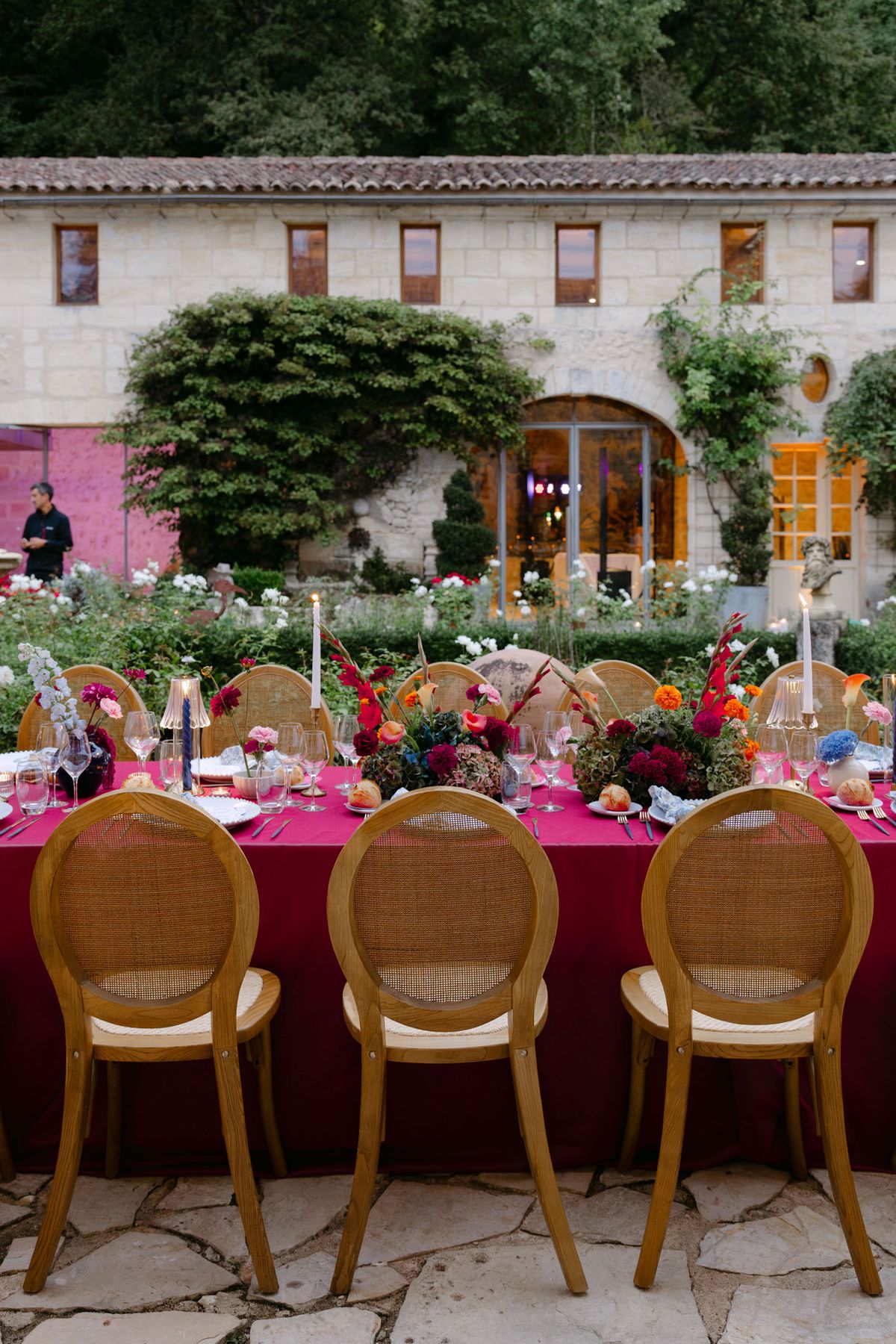 Outdoor reception in the stone patio courtyard at Domaine de Perrotin, with long tables dressed in raspberry-red linen, gold cane chairs and colourful florals for Rooa and Mark's wedding