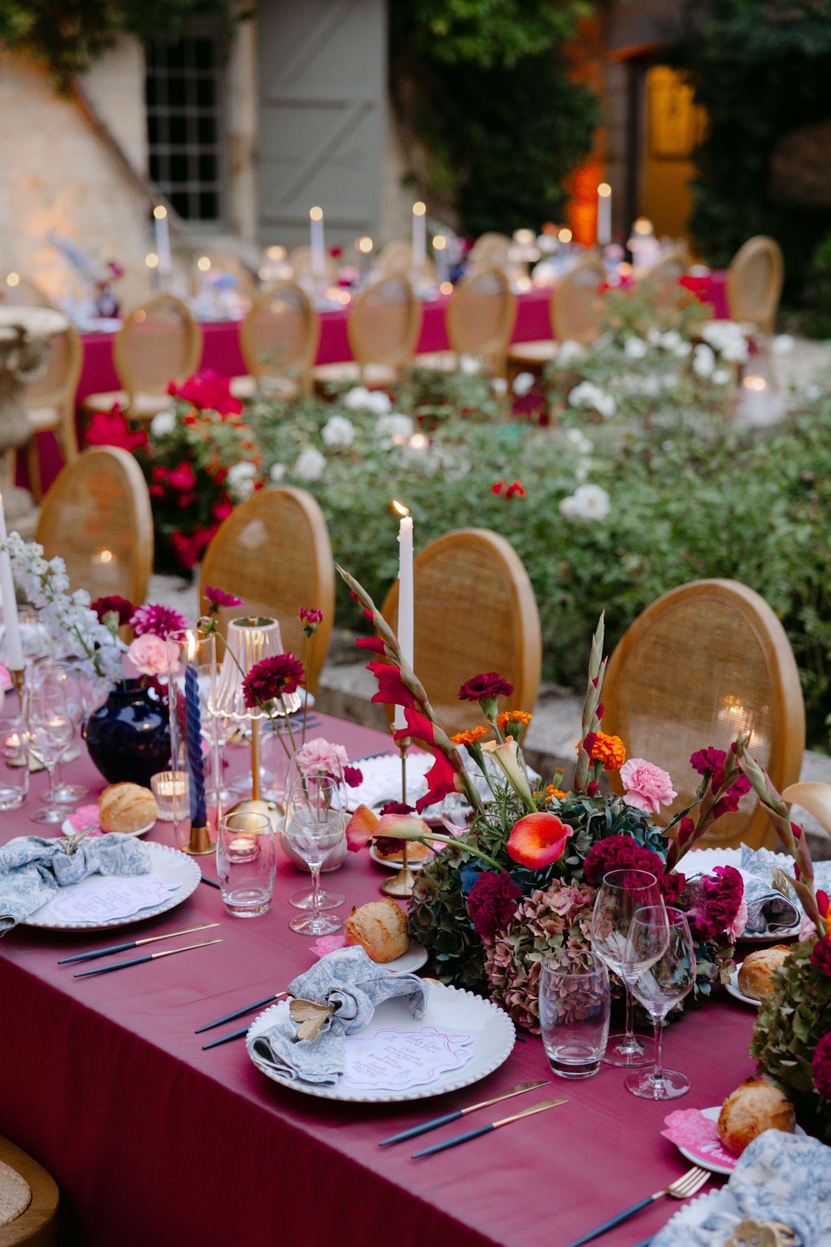 Outdoor reception in the stone patio courtyard at Domaine de Perrotin, with long tables dressed in raspberry-red linen, gold cane chairs and colourful florals for Rooa and Mark's wedding