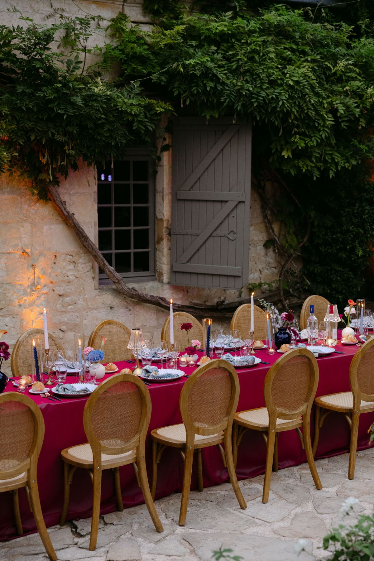 Outdoor reception in the stone patio courtyard at Domaine de Perrotin, with long tables dressed in raspberry-red linen, gold cane chairs and colourful florals for Rooa and Mark's wedding