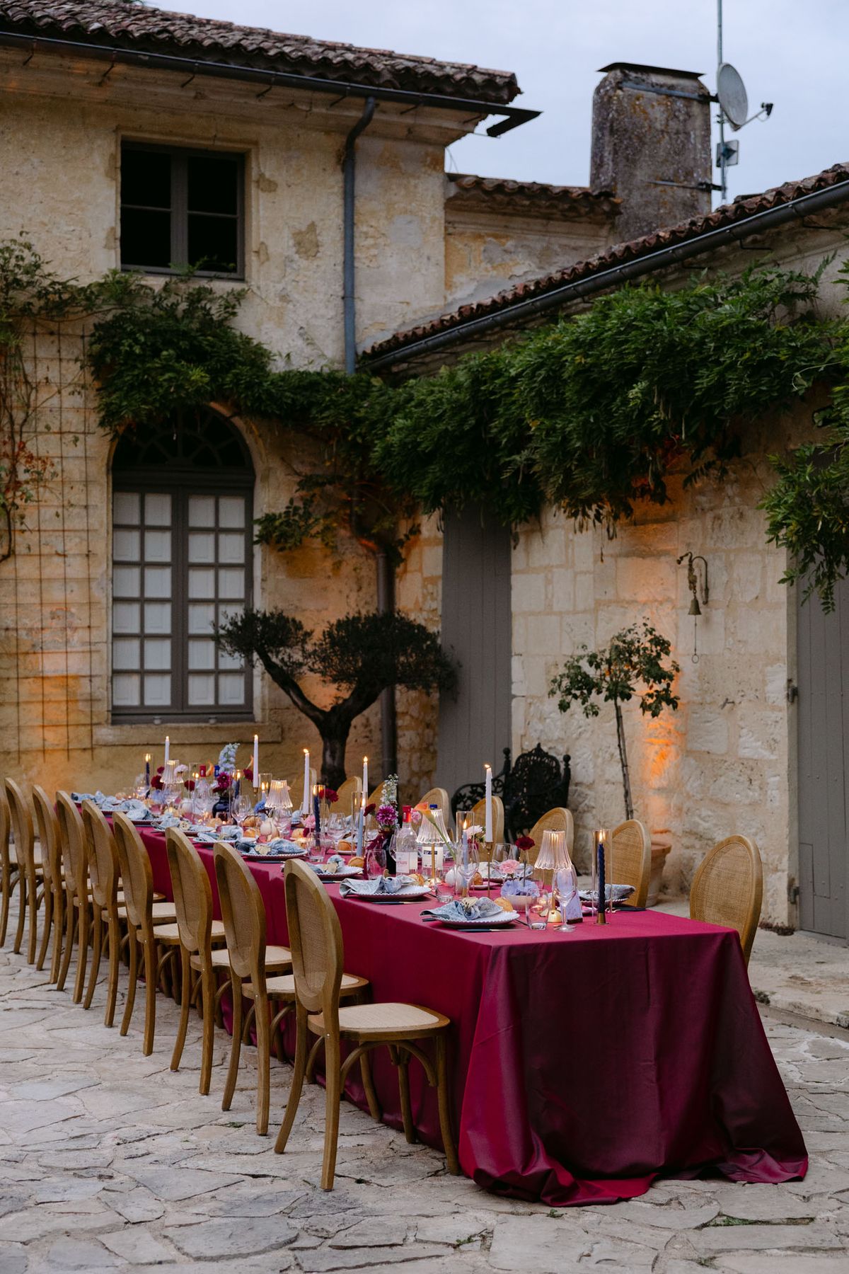 Outdoor reception in the stone patio courtyard at Domaine de Perrotin, with long tables dressed in raspberry-red linen, gold cane chairs and colourful florals for Rooa and Mark's wedding