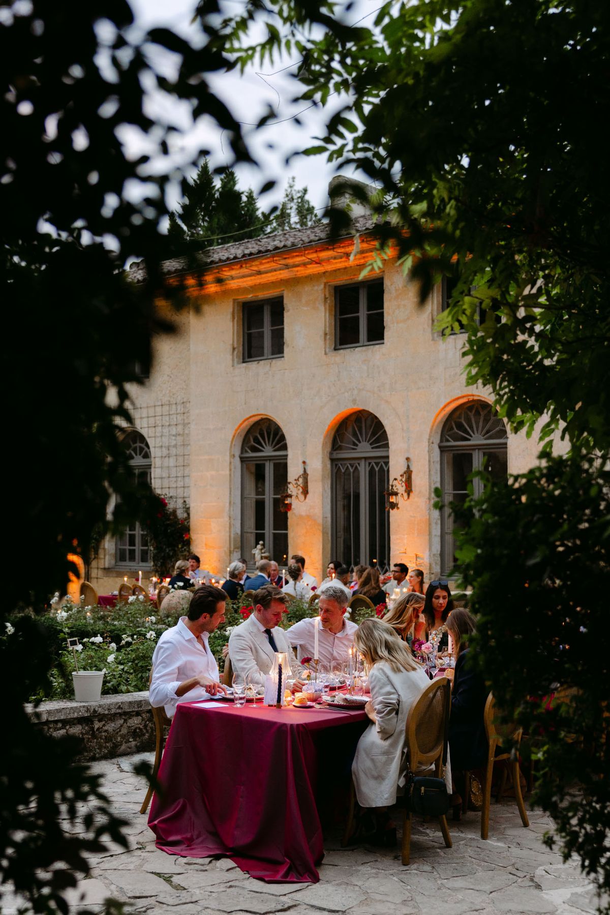 Outdoor reception in the stone patio courtyard at Domaine de Perrotin, with long tables dressed in raspberry-red linen, gold cane chairs and colourful florals for Rooa and Mark's wedding